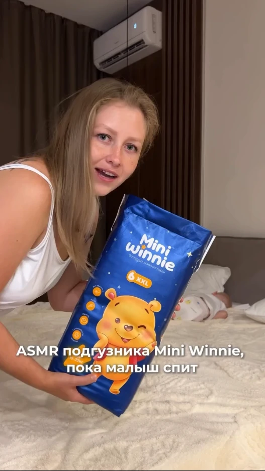 Babae na may hawak na isang pakete ng Mini Winnie diapers malapit sa isang natutulog na sanggol sa isang kama.